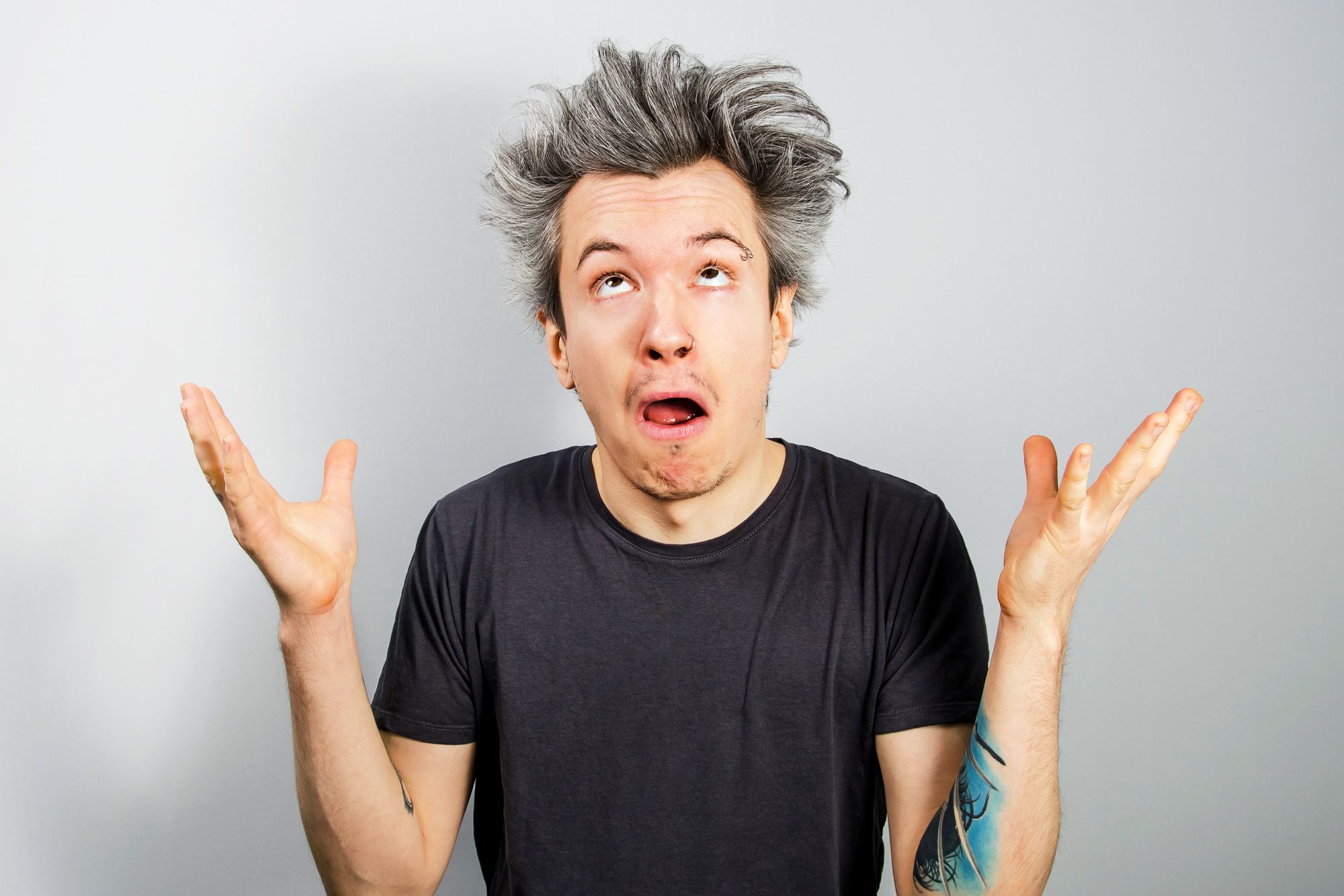 Unshorn and unshaven young person with piercings on their face, shrugs his shoulders and looking up, on gray background.