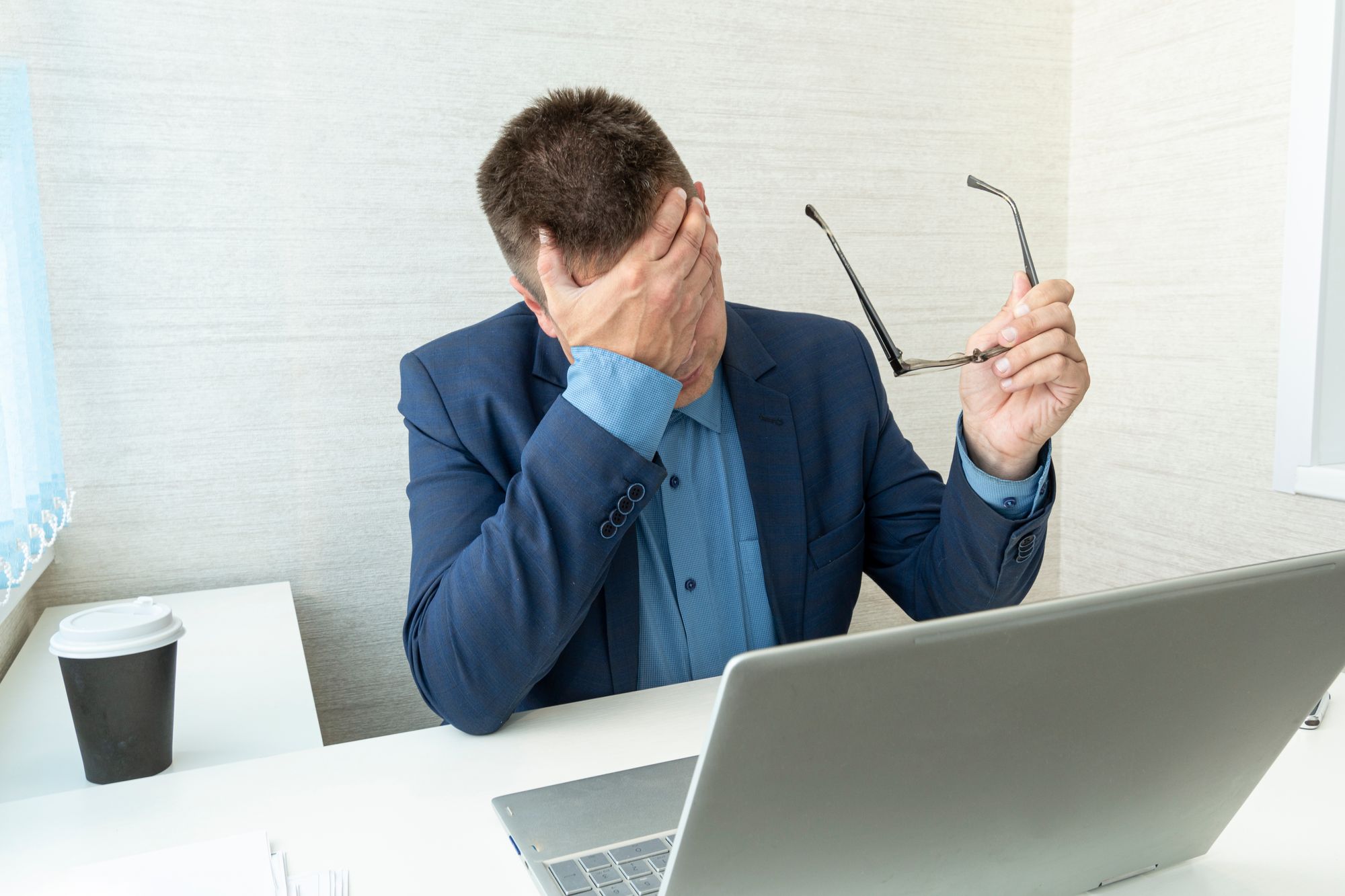 An office worker in front of a laptop, looking worried, taking off their glasses and putting one hand over their face.