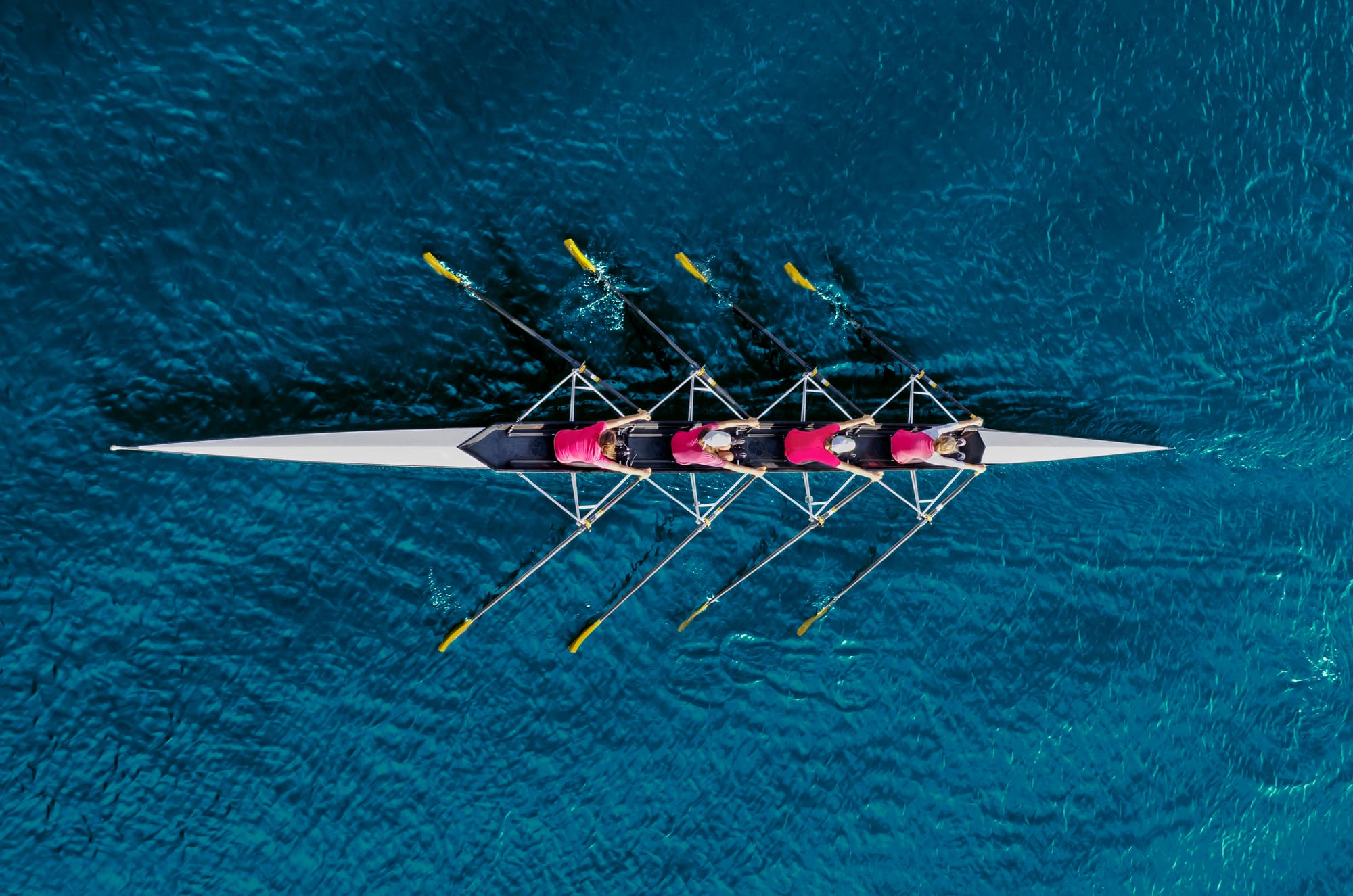  Aerial view of four people propelling a rowboat through water.