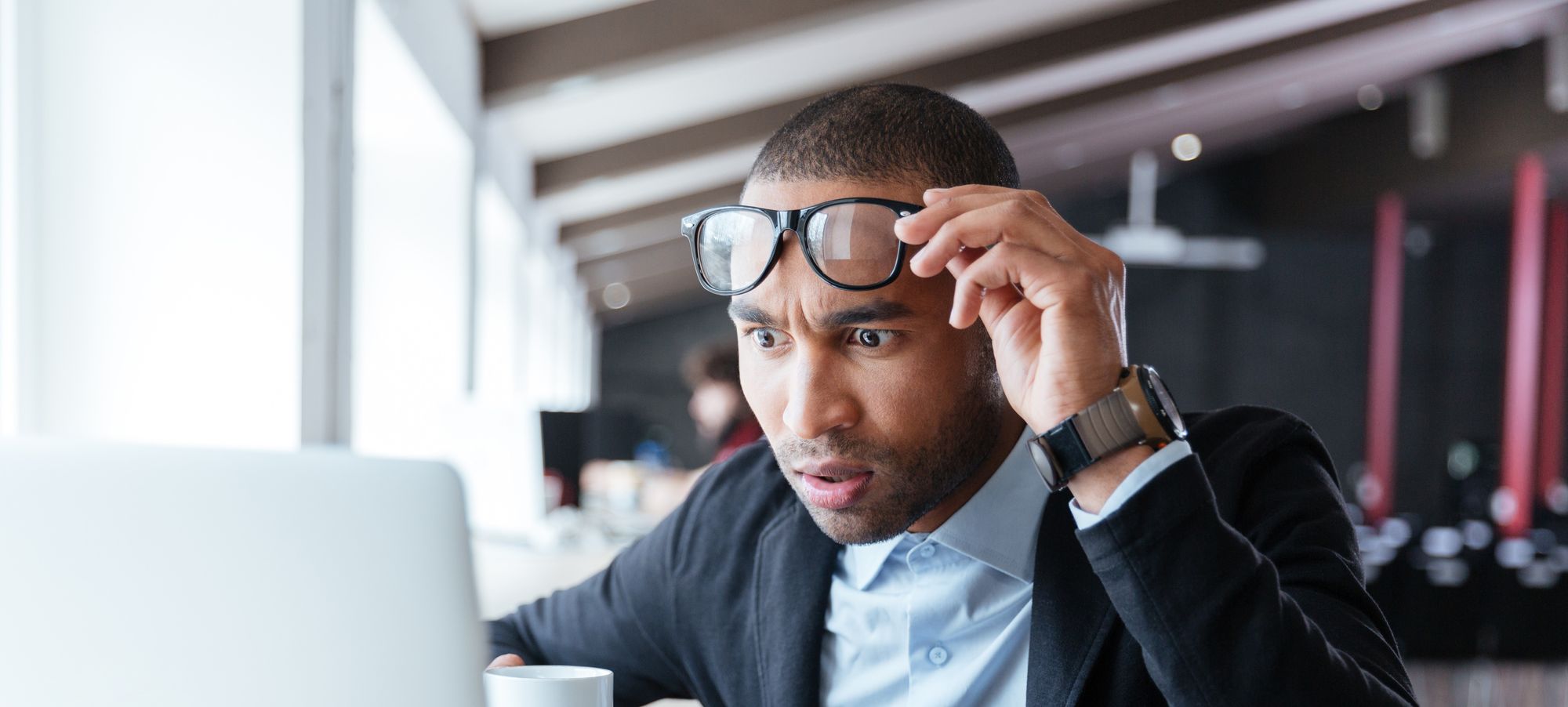 A person looking at a laptop holding their glasses above their eyes with a concerned look on their face.